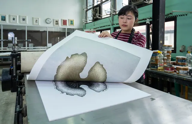 A woman inspects a freshly printed design in a modern printmaking studio, surrounded by printing tools and equipment. Anthotype Process with Spinach Juice: Sun-Printed Photographs
